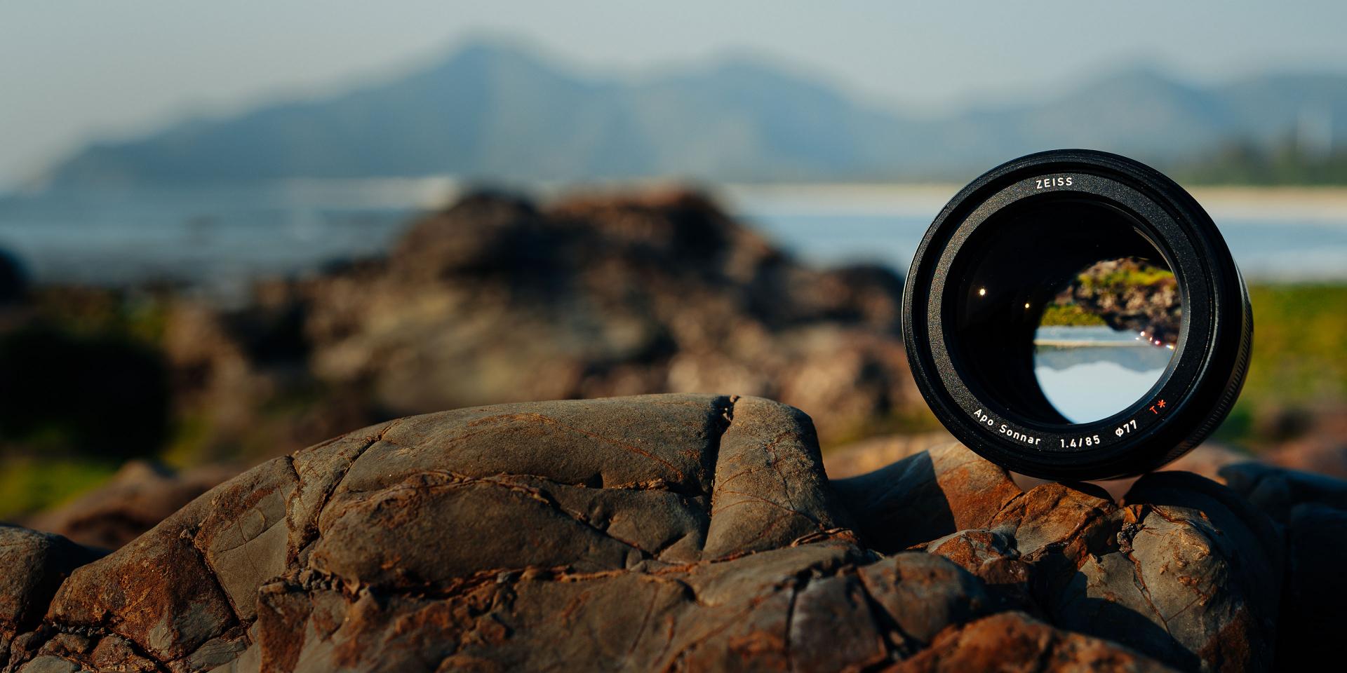 A ZEISS camera lens is placed on a rock with a scenic outdoor background of mountains and a blurred body of water.