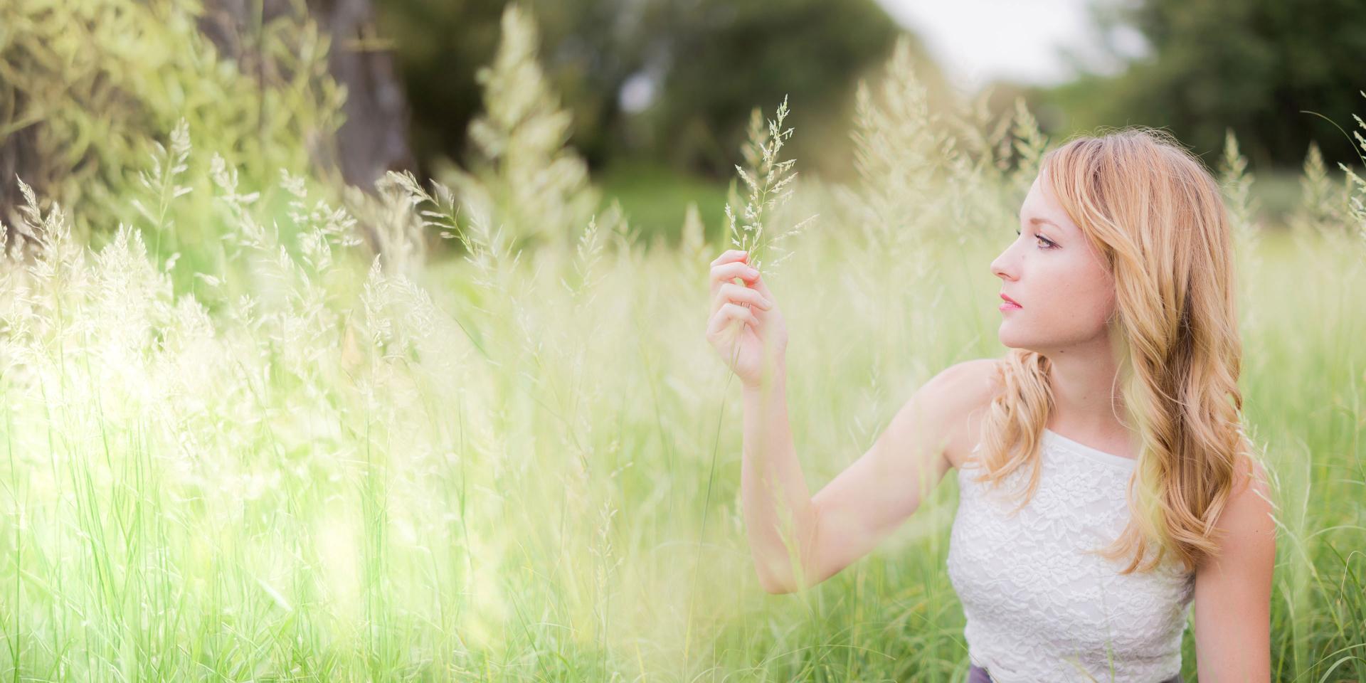 A serene portrait of a woman in a sunlit field, gently touching tall grass, showcasing the natural colors and fine details captured by ZEISS optics.