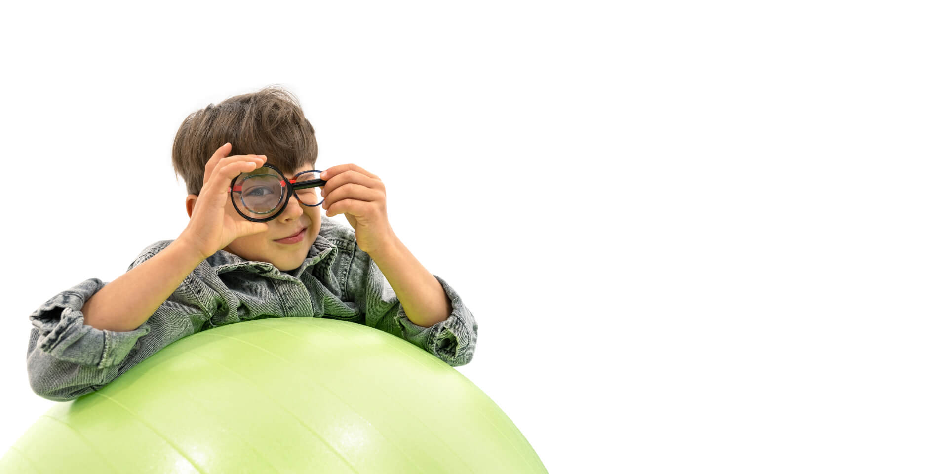 男孩戴着蔡司近视眼管理镜片，靠在健身球上，将放大镜放到一只眼睛前。