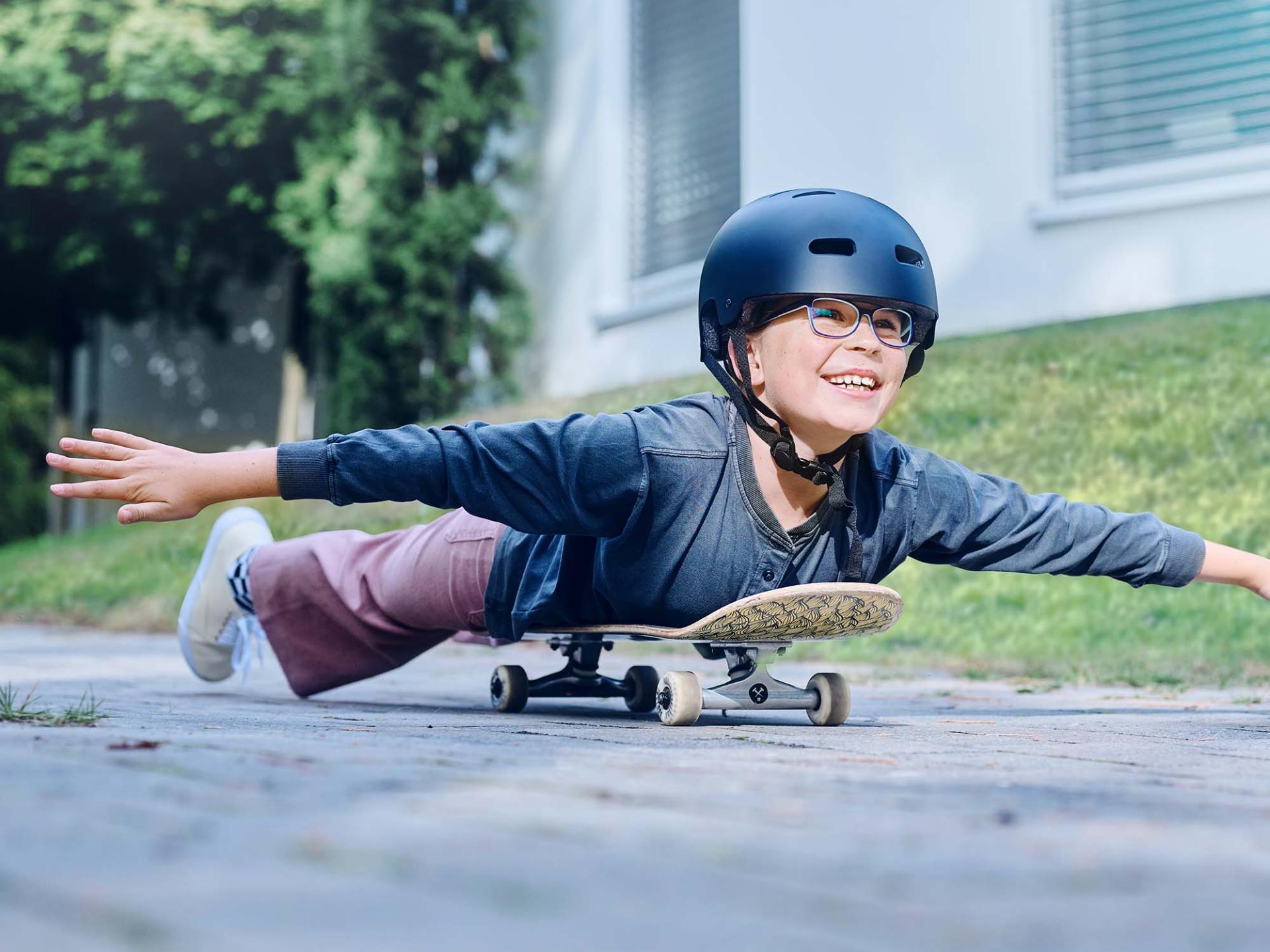女孩戴着头盔和眼镜，躺在滑板上滑下马路，伸开双臂。